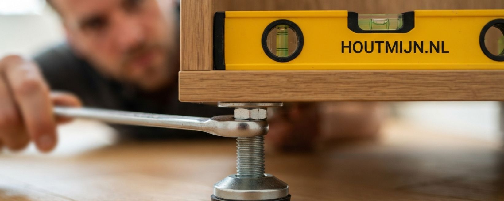 Een close-up op ooghoogte van een man die, met onscherpe achtergrond, geconcentreerd een in hoogte verstelbare houten meubelpoot stelt met behulp van een steeksleutel. Op het meubel rust een geel waterpas met de zwarte opdruk HOUTMIJN.NL.
